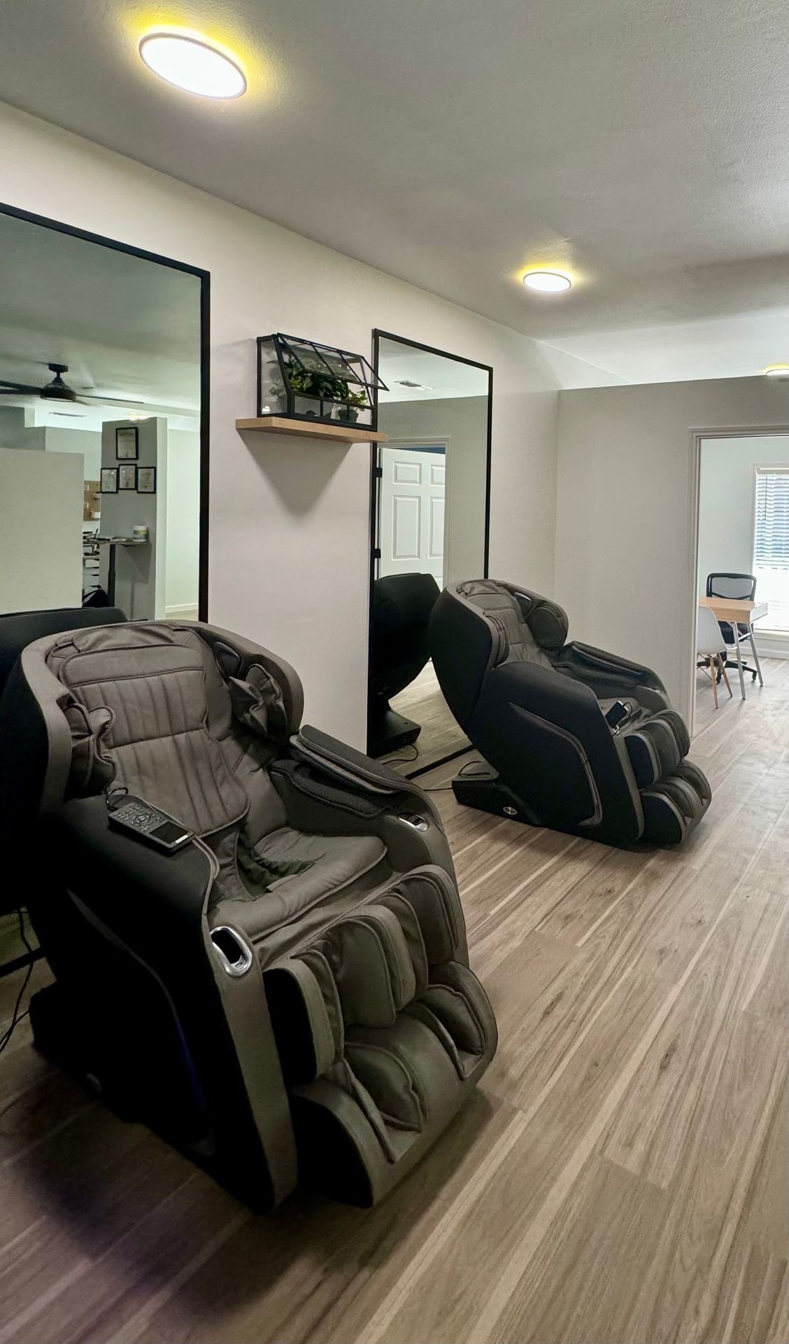 Room with two massage chairs, wood flooring, and a shelf with a plant under ceiling lights.